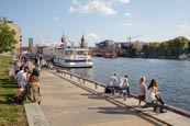 People Relaxing By The River Spree Near Oberbaumbrücke, In The Former Berlin Wall Death Strip, Berli