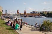 People Relaxing By The River Spree Near Oberbaumbrücke, In The Former Berlin Wall Death Strip, Berli